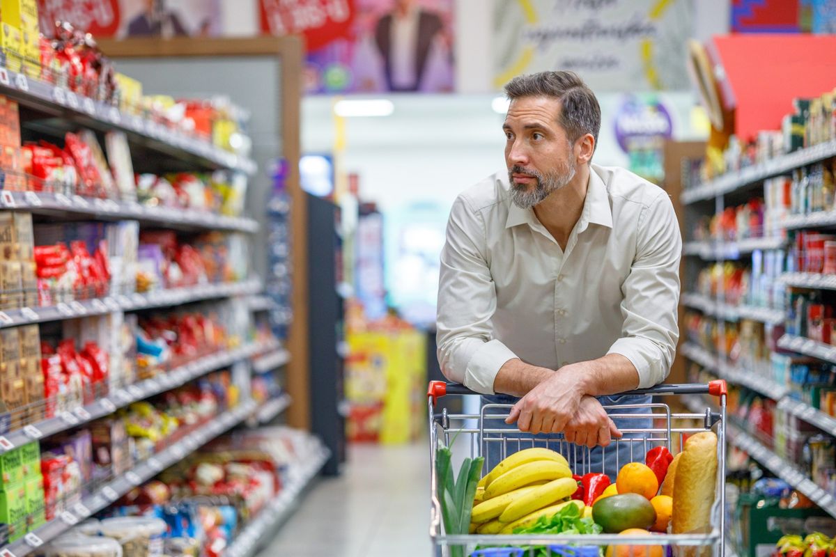 A man leaning forward on a grocery cart in a supermarket aisle, forearms resting on the handle.