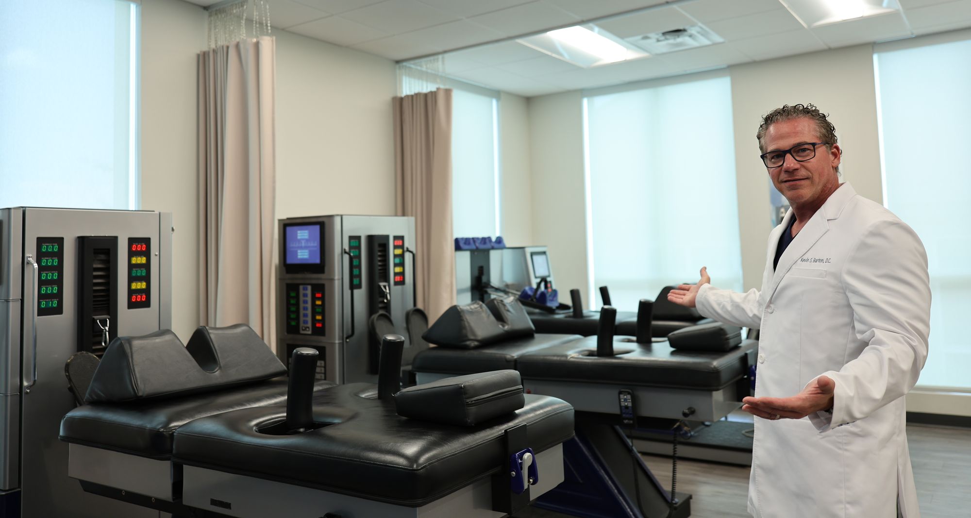Dr. Kevin Barton in his white coat, gesturing toward the DRX9000 spinal decompression tables in the Texas Spine Clinic treatment room.
