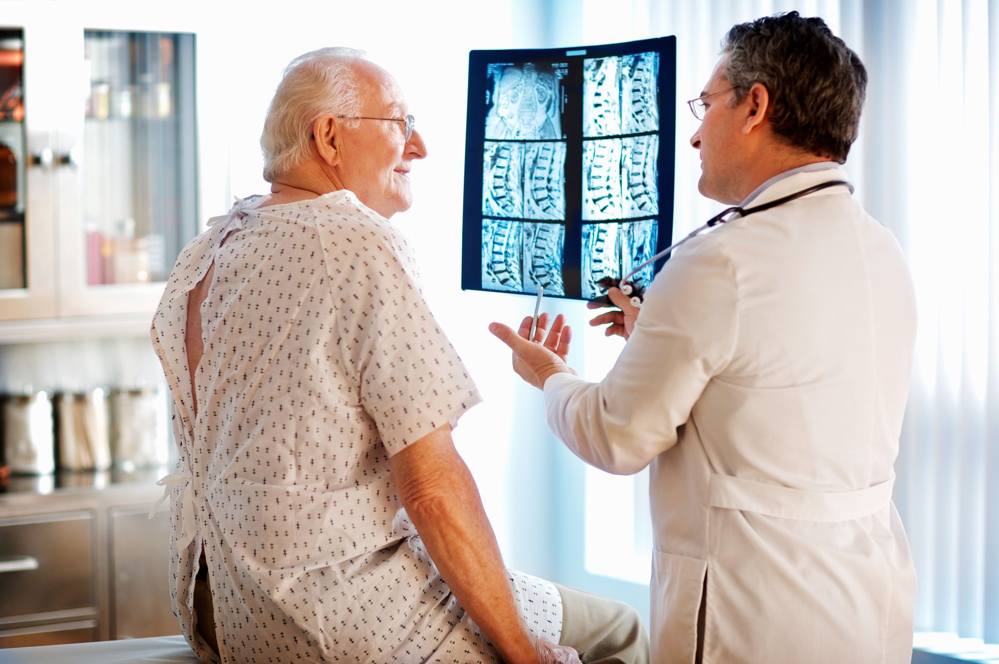 A doctor holding up spine films and going through them with an older patient in a gown.
