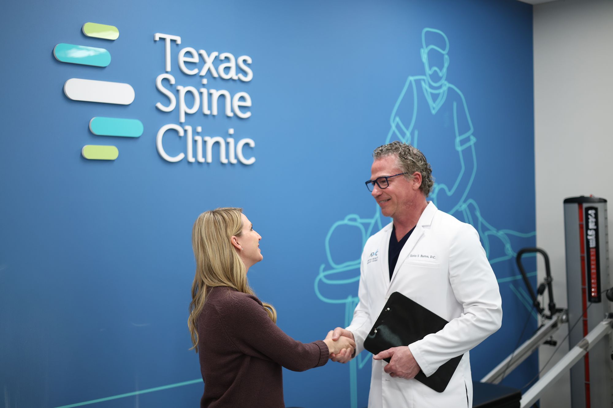 A Texas Spine Clinic doctor greeting a patient at the clinic's logo wall.