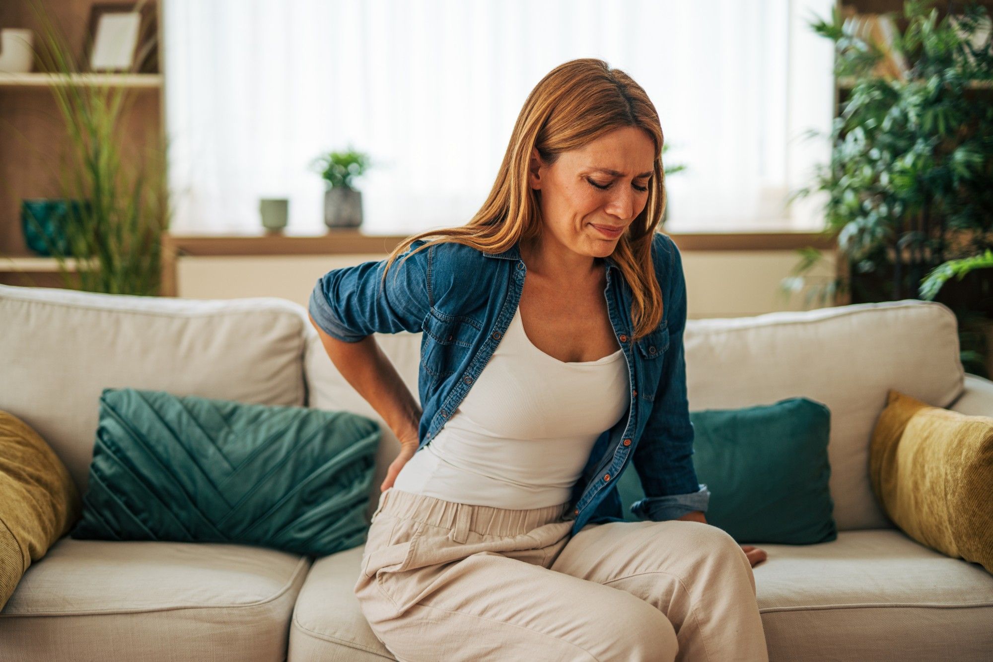 A woman sitting on her couch, wincing and holding her lower back.