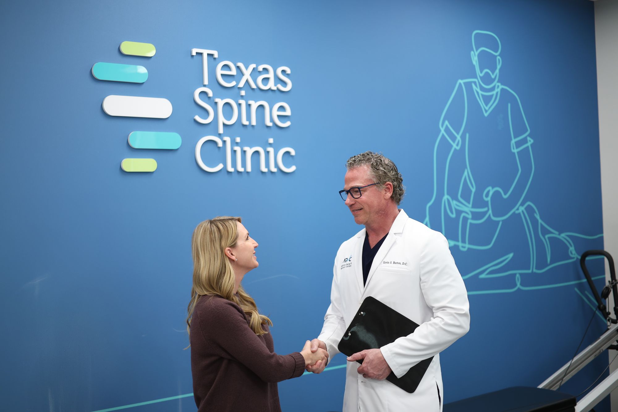 Dr. Barton shaking hands with a new patient in front of the Texas Spine Clinic logo wall.