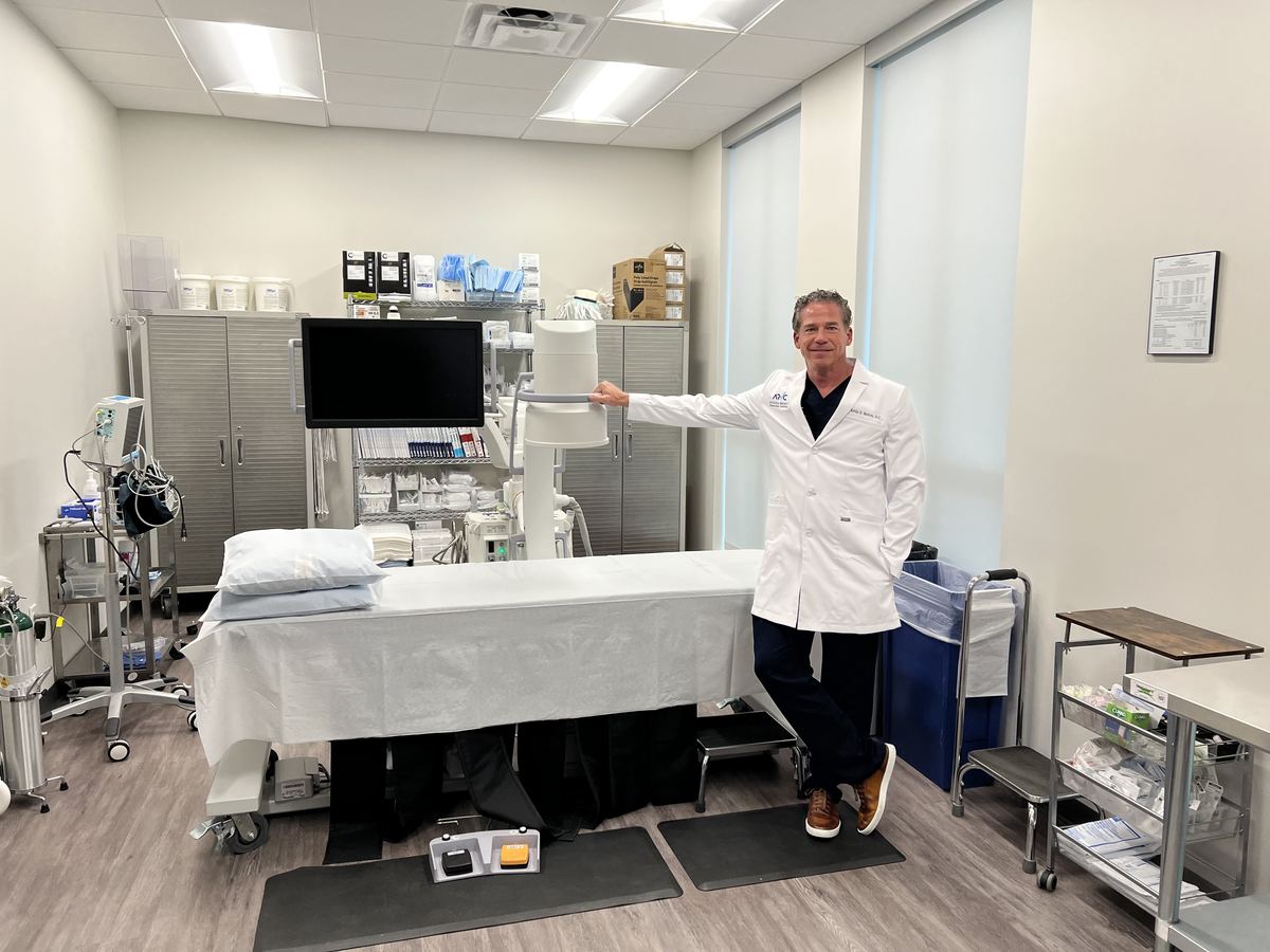 Dr. Kevin S. Barton standing beside the procedure table in the Texas Spine Clinic procedure room, with an imaging unit and monitor.