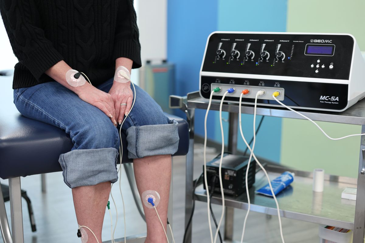 A patient seated beside the scrambler therapy machine with electrode pads on the backs of both wrists and on both shins, the leads running to the machine on its cart.
