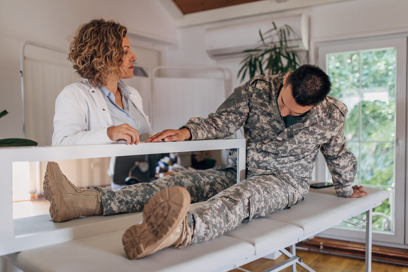 Stock illustration: a soldier seated on a treatment table with a therapy mirror positioned so his intact leg's reflection appears in place of the missing one, a clinician beside him.