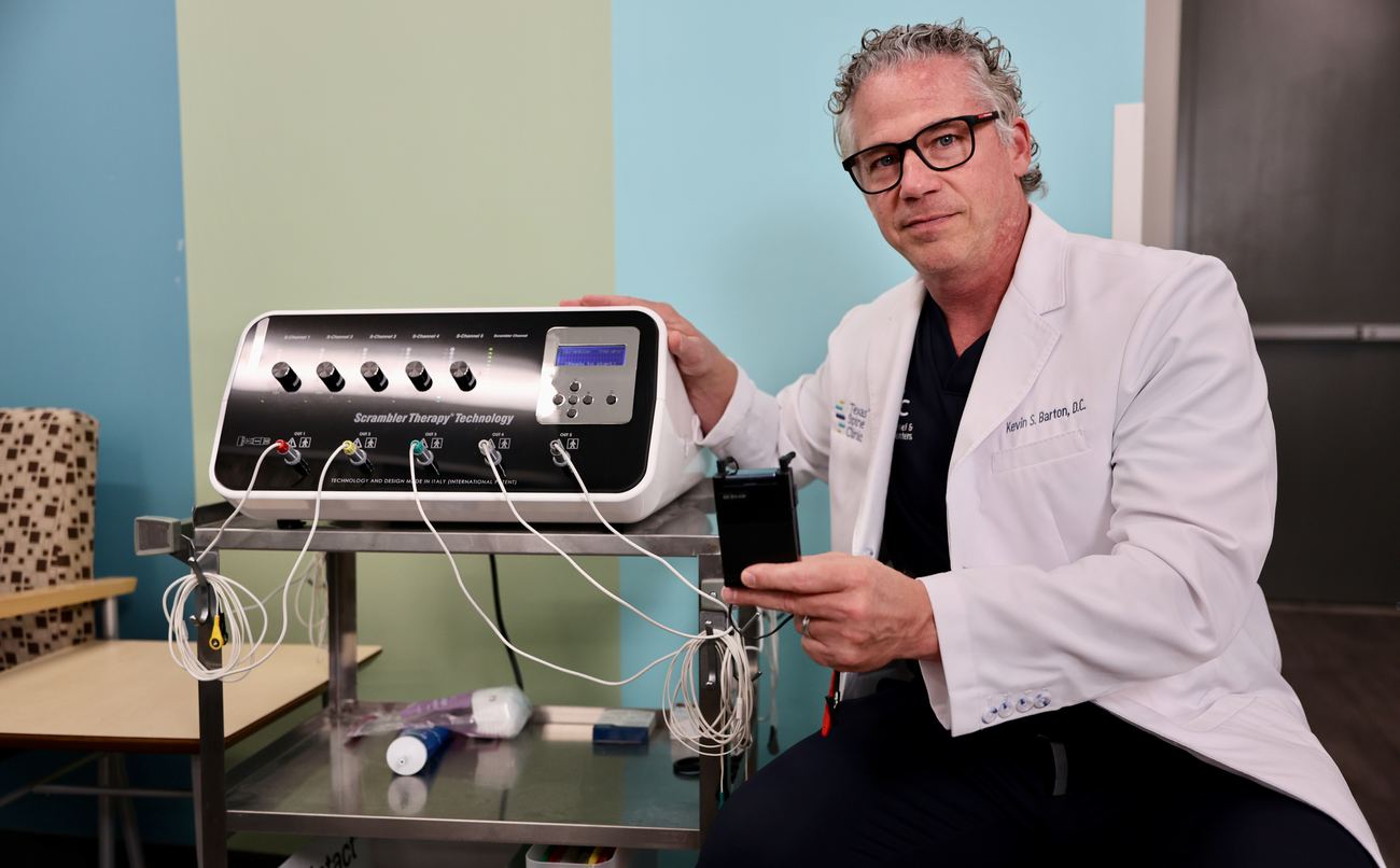 Dr. Barton with one hand on the ST-5A scrambler therapy machine, holding a pocket-sized TENS unit in his left hand for scale.